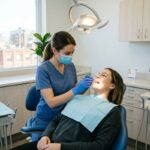 Dentist examines a patient's open mouth using dental tools in a clinic chair under an overhead light.